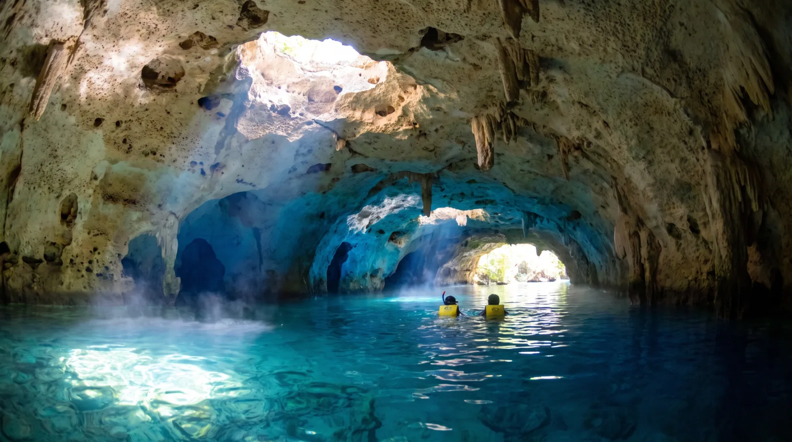 Dos personas con chaleco salvavidas nadando por una caverna de piedra caliza con agua turquesa y haces de luz